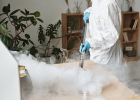Woman in protective gear disinfects a wooden table, indoor setting, using fumigation method.